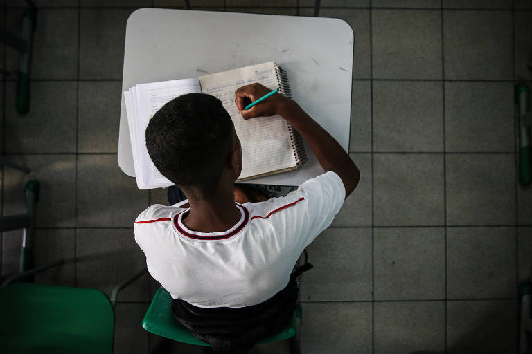 Vista de cima, a imagem mostra um estudante sentado em uma cadeira verde, concentrado em ler e fazer anotações em seu caderno, que está sobre uma mesa branca. O aluno veste um uniforme escolar branco com detalhes em vermelho e preto, sugerindo um ambiente de aprendizado formal.