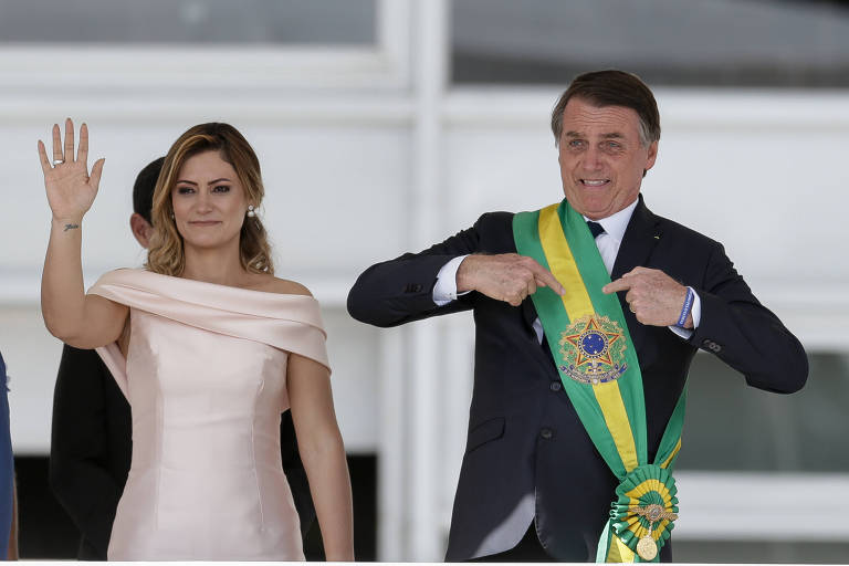 Jair Bolsonaro, ao lado da esposa, aponta com as duas mãos para a faixa presidencial verde-amarela em seu peito  