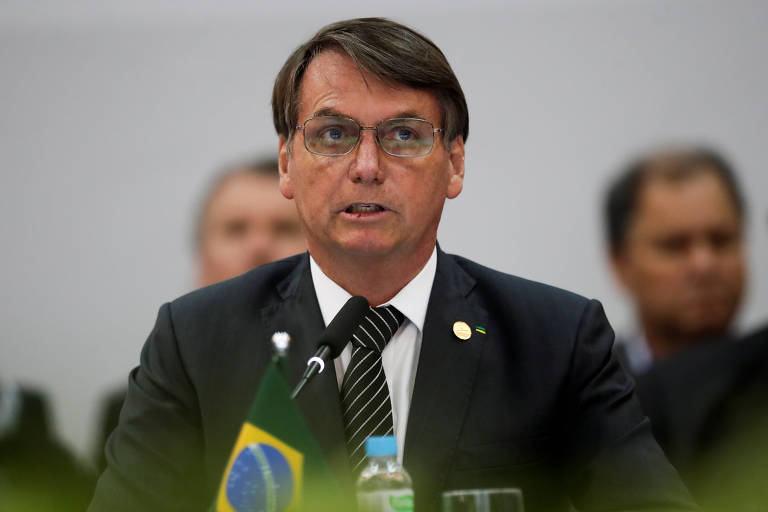 O presidente Jair Bolsonaro aparecente em frente a um microfone, com uma pequena bandeira do Brasil em sua frente