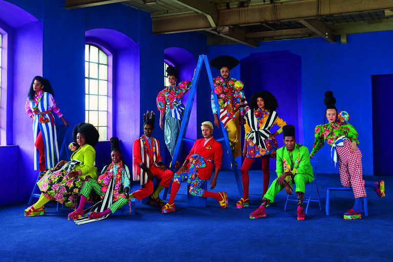 Onze pessoas com roupas coloridas de neón posam lado a lado em um salão cujas paredes estão iluminadas de roxo