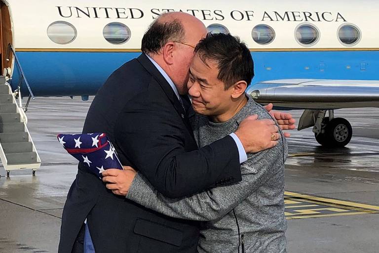 O sino-americano, vestido com um moletom cinza, abraça o embaixador americano enquanto segura uma bandeira dos Estados Unidos enrolada. Atrás deles, um  avião oficial em que se lê "Estados Unidos da América", em inglês. 