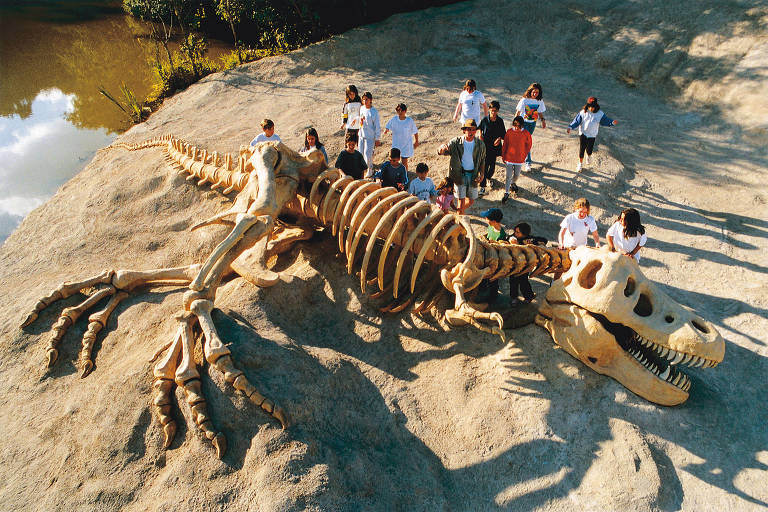 Réplica de fóssil de dinossauro deitado na areia, um grupo de crianças está na parte superior dele.