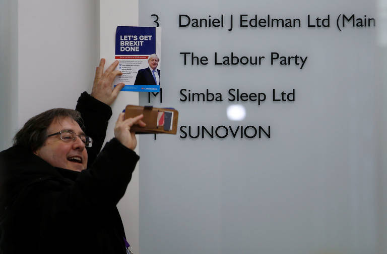 Homem segura um folheto do primeiro-ministro Boris Johnson ao lado da placa do Partido Trabalhista dentro do prédio que abriga a sede do partido, após a eleição geral em Londres