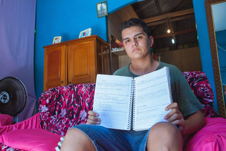 Um adolescente está sentado em uma cama e segura um caderno escolar