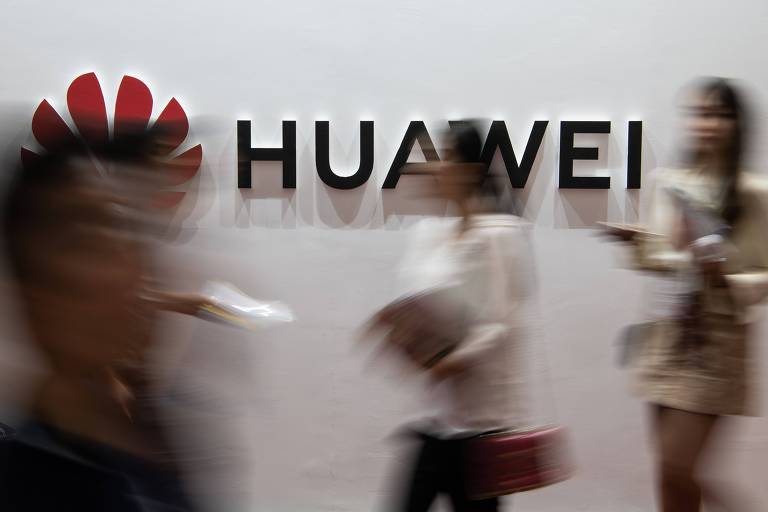 Pessoas transitam em frente a símbolo da Huawei colocado em parede branca