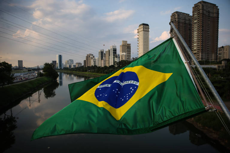Vista de prédios de alto padrão na marginal Pinheiros, com bandeira do Brasil à frente pendurada na ponte Cidade Jardim