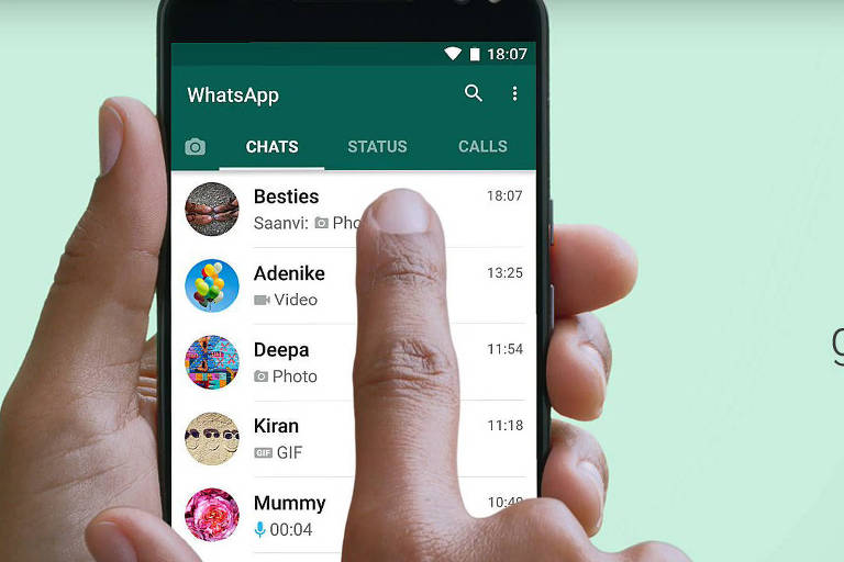 Telefone celular contra fundo verde é segurado nas mãos de pessoa de pele clara, com o aplicativo WhatsApp aberto, mostrando contatos