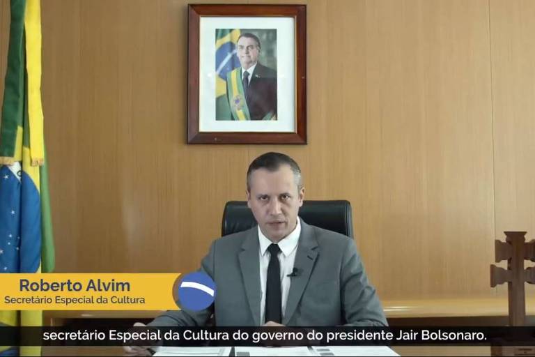 Imagem mostra Roberto Alvim ao centro, sentado em uma mesa; acima dele, a foto do presidente Jair Bolsonaro; ao seus lados, a bandeira brasileira e uma cruz