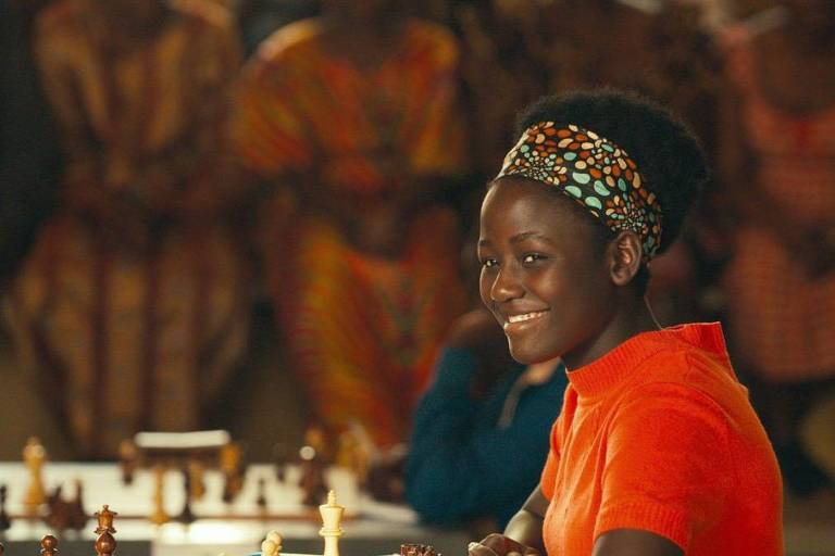 Cena do filme "Rainha de Katwe". Na foto aparece uma mulher negra jovem. Ela sorri e usa uma camiseta laranja e um turbante com detalhes verdes e avermelhados no cabelo crespo. Ela está sentada em uma mesa com tabuleiro e peças de xadrez, no fundo da imagem é possível ver outras pessoas no ambiente. Isso sugere que a protagonista do filme está em um torneio. 