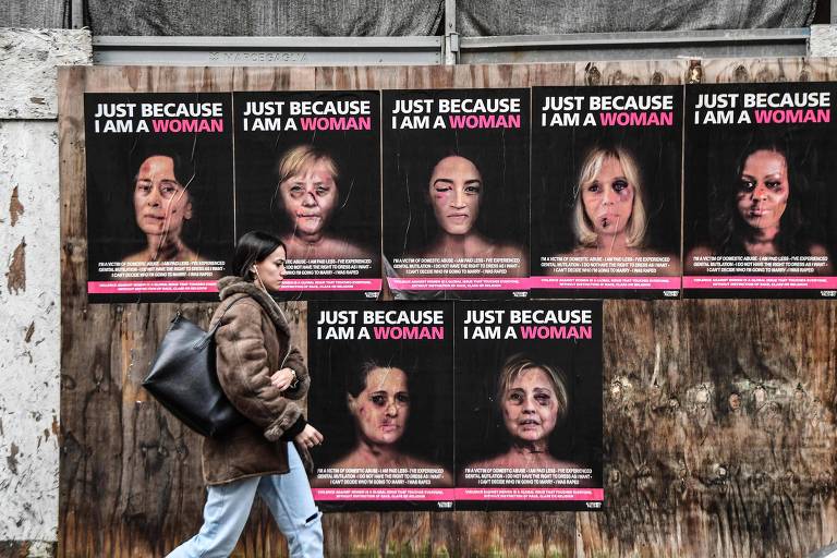 Pedestre caminha em frente a cartazes com imagens de protagonistas do âmbito político retratadas como vítimas de agressão, em Milão; da esquerda para a direita, do alto, estão representadas Aung San SuuKyi, Angela Merkel, Alexandria Ocasio-Cortez, Brigitte Macron, Michelle Obama, Sonia Gandhi e Hillary Clinton