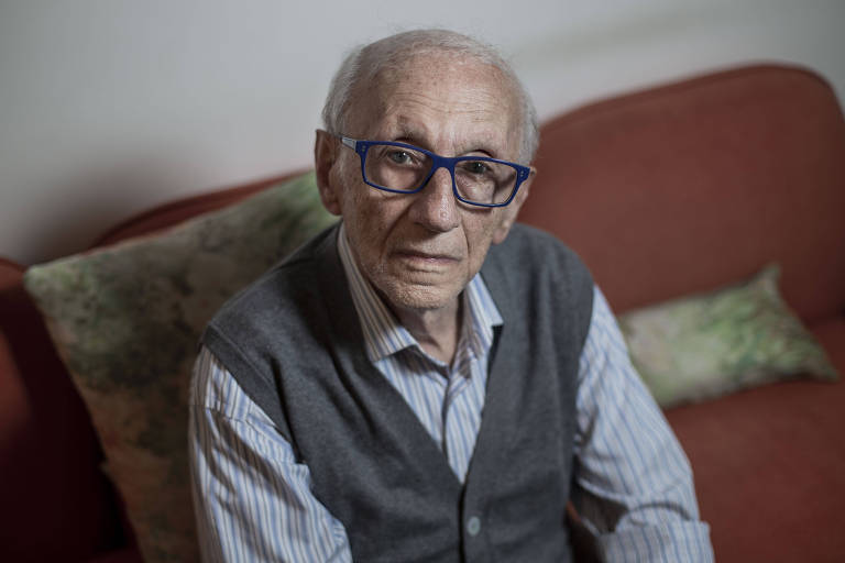Imagem em primeiro plano mostra homem idoso de óculos sentado posando para foto