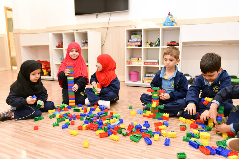 Três meninas com véu islâmico e dois meninos com uniforme. Os cinco brincam de lego sentados no chão