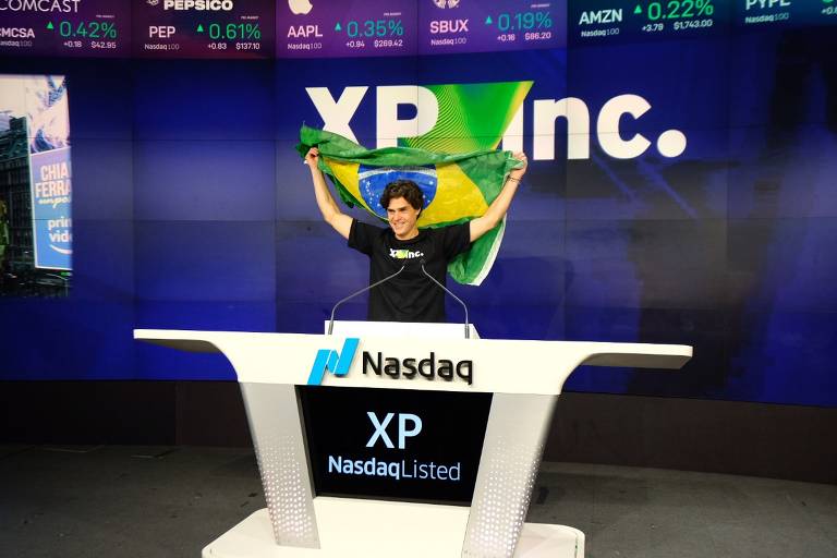 O fundador da XP, Guilherme Benchimol, posa atrás da bancada da Nasdaq segurando uma bandeira do Brasil no alto. Atrás, o logo da XP Inc. aparece escrito em um fundo azul escuro