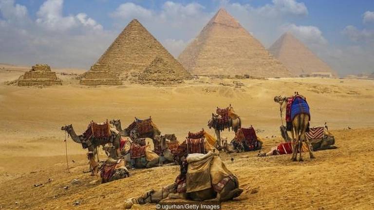 Camelos com pirâmides ao fundo, em deserto 