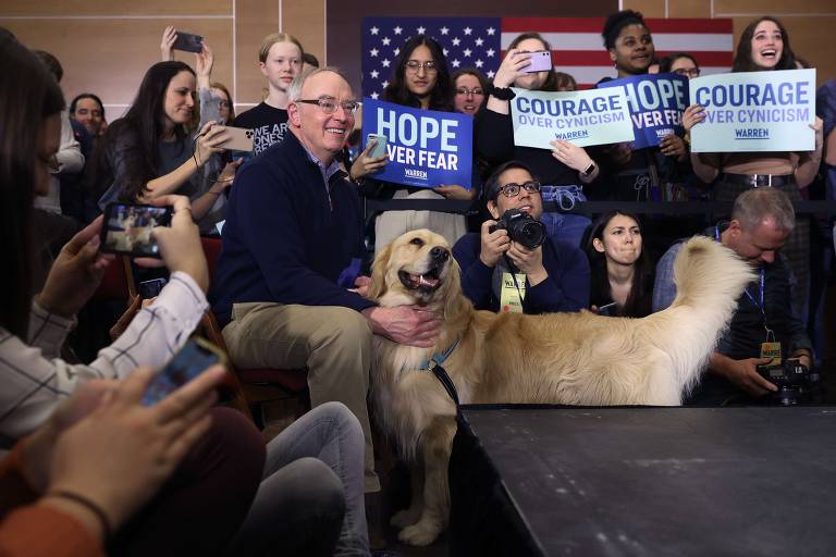 Enquanto Elizabeth Warren discursa, apoiadores exibem cartazes e Bruce Mann, seu marido, posa com cachorro; evento aconteceu no campus da Universidade de Simpson, em Indianola, Iowa