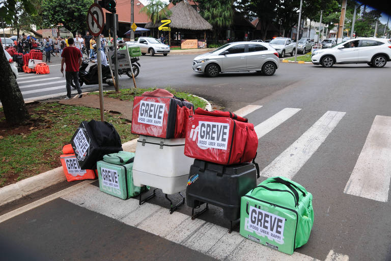 Retrato de mochilas de entregadores depositadas na faixa de pedestres de uma avenida; nelas, estão escritas "greve"