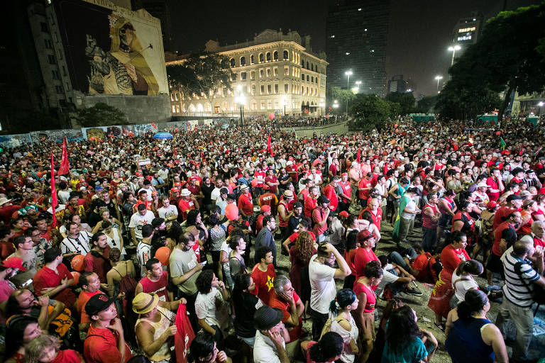 A imagem mostra uma grande multidão reunida em um espaço público à noite, com muitas pessoas vestindo roupas vermelhas. Ao fundo, há um edifício iluminado e uma grande muralha com uma arte visível. A atmosfera parece ser de protesto, com bandeiras vermelhas visíveis entre a multidão.