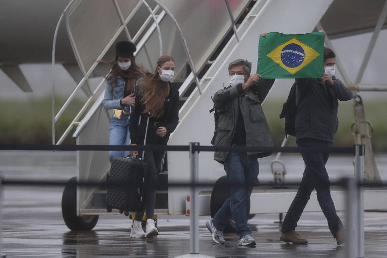 Quatro pessoas descem de um avião， usando máscaras. Uma delas segura uma bandeira do Brasil. O ambiente parece ser um aeroporto， com uma escada de desembarque ao fundo e um dia nublado.