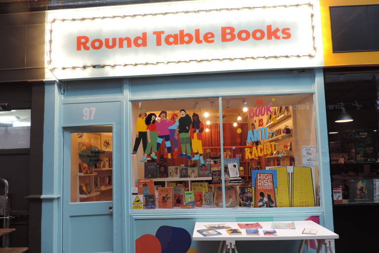 Vitrine de livraria vista da rua. Tem faixa branca com nome Round Table Books escrito em vermelho. Paredes da fachada azuis. Ambiente iluminado em tom amarelo dentro, com livros e outros artigos pendurados e organizados em prateleiras.