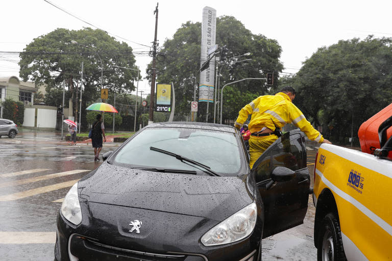 Homem trabalha em um carro danificado em uma rua inundada após fortes chuvas no bairro do Butantã, em São Paulo