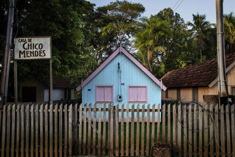 Casa de madeira azul com janelas rosa claro; ao lado há uma placa dizendo "casa de Chico Mendes"