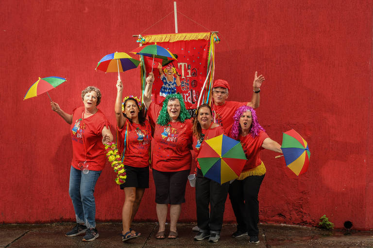 Cinco mulheres e um homem, uniformizados com camisetas com o logo do bloco Tia eh o Caraleo, segurando guarda-chuvas de frevo e um estandarte do bloco, fazendo caretas para a câmera