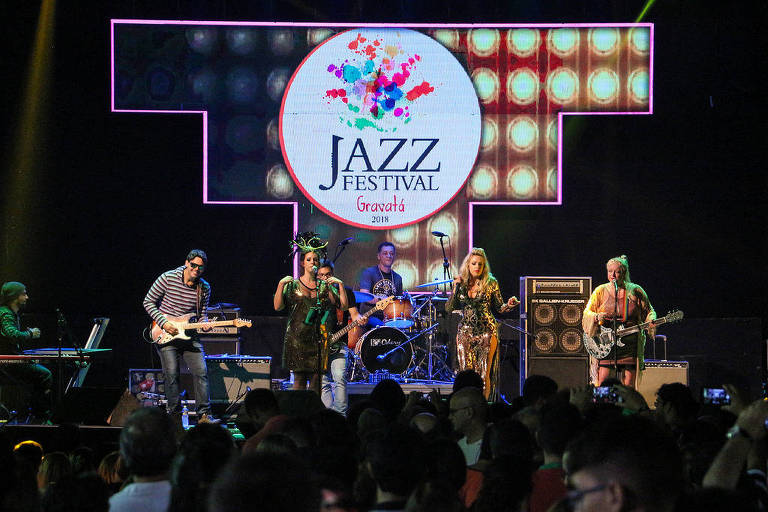 Banda de jazz com guitarrista, baixista, duas vocalistas e baterista toca em palco iluminado