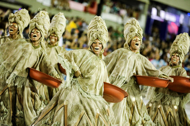 a foto mostra mulheres que desfilam caracterizadas de ganhadeiras, com roupa e maquiagem dourada, durante um desfile de carnaval