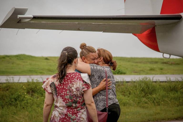 Três pessoas se abraçam enquanto choram próximas a um avião de pequeno porte