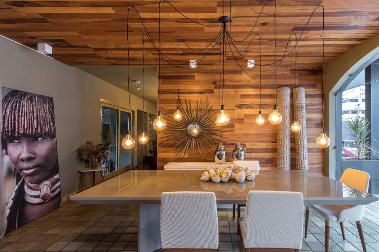 Sala de jantar com parede e teto de madeira, mesa de madeira quadrada, cadeiras brancas e quadro de indígena apoiado no chão 