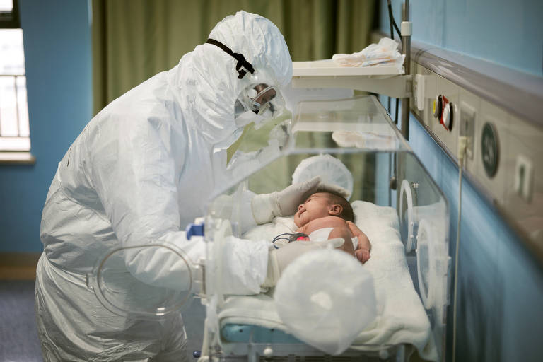 De roupa especial e máscara,  médico mexe em bebê em berço em hospital