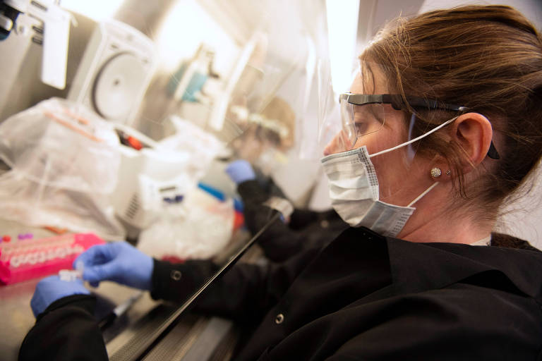Pesquisadora com máscara que cobre nariz e boca, óculos de proteção transparente com aro preto, roupa preta e luvas azuis manipula pequeno recipiente em ambiente de laboratório