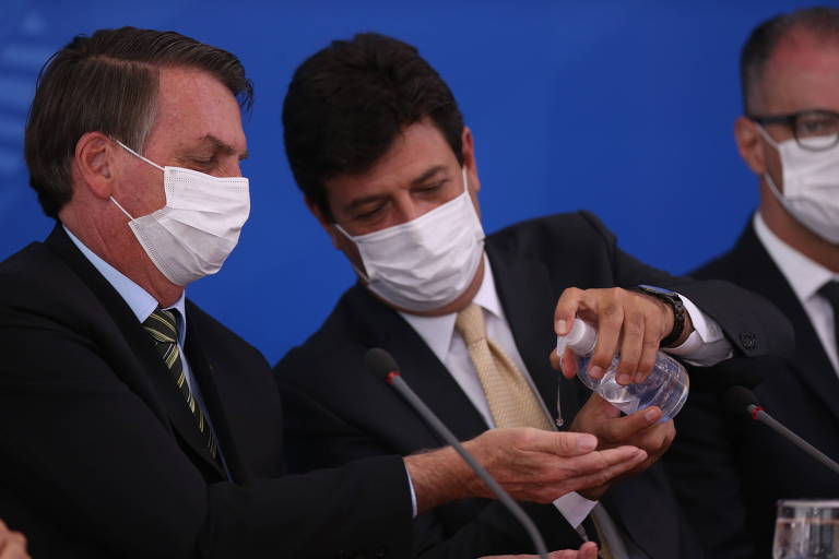 O ministro da Saúde, Luiz Henrique Mandetta (à dir.), passa álcool gel na mão do presidente Jair Bolsonaro, ambos de máscara, durante coletiva de imprensa para falar sobre a crise do coronavírus