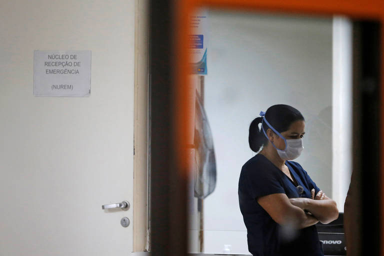 Ao lado de uma porta aberta, enfermeira usando máscara cirúrgica e uniforme está de braços cruzados