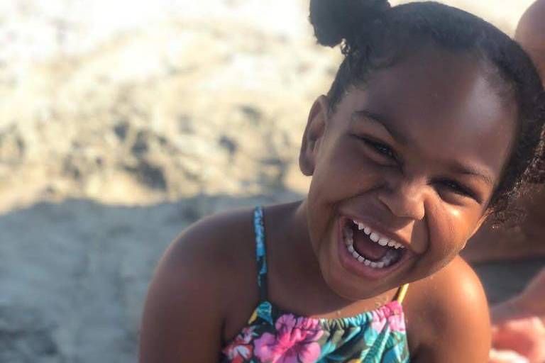 Menina de 4 anos chamada Catarina na praia. Usa duas chuquinhas altas na cabeça. Ela é negra com sorriso largo. Usa biquini florido