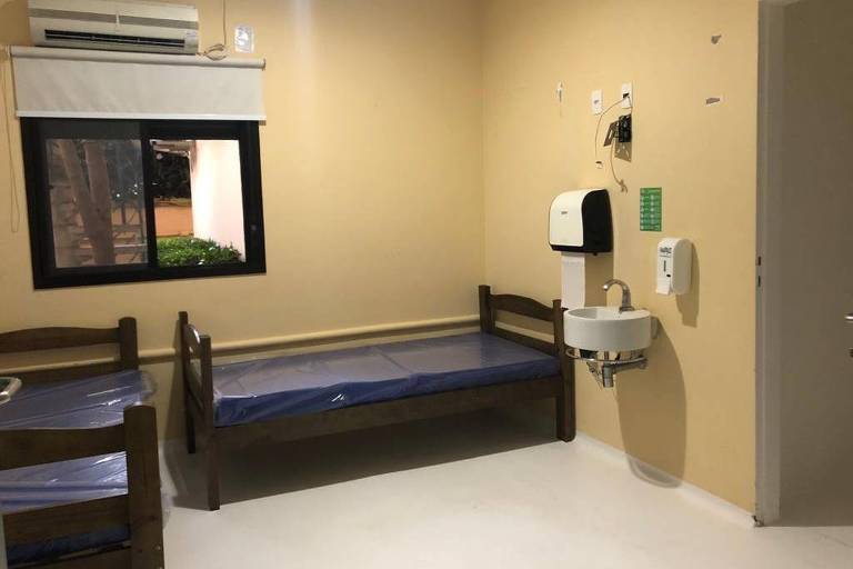 Quarto de hospital com duas camas de solteiro, uma pia, uma janela com persiana e ar condicionado