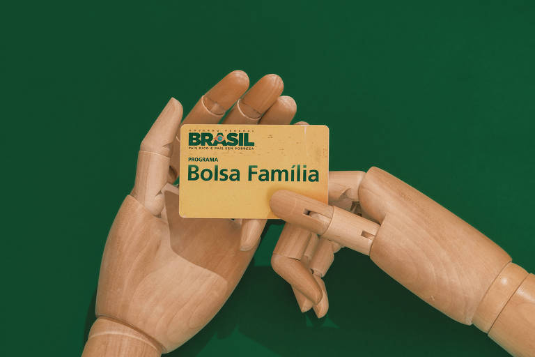 Foto mostra um fundo verde com duas mãos articuladas de madeira segurando um cartão amarelo escrito Bolsa Família em verde.