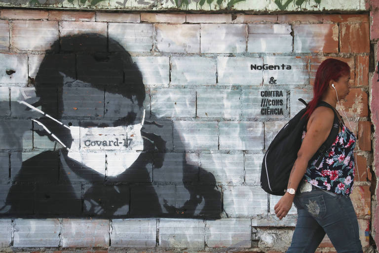 Uma mulher caminha em frente a um muro com um graffiti representando Jair Bolsonaro; o presidente usa uma máscara que está escrito 'covard-17'