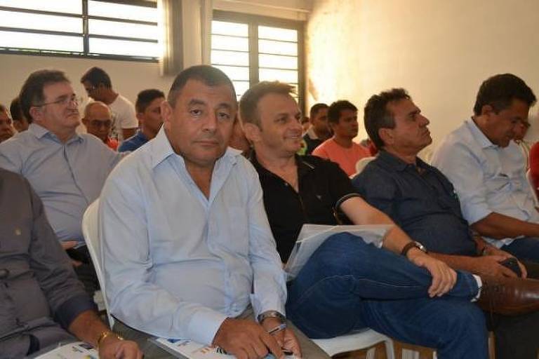 De camisa azul de manga cumprida, Antônio Felícia aparece em sala com janela e porta ao fundo. Ele está sentado com as mãos sobre o colo.