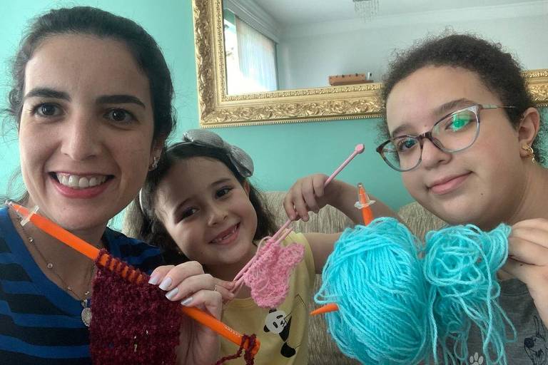 Mãe e as duas filhas seguram novelos de lã coloridos e agulhar de tricô 