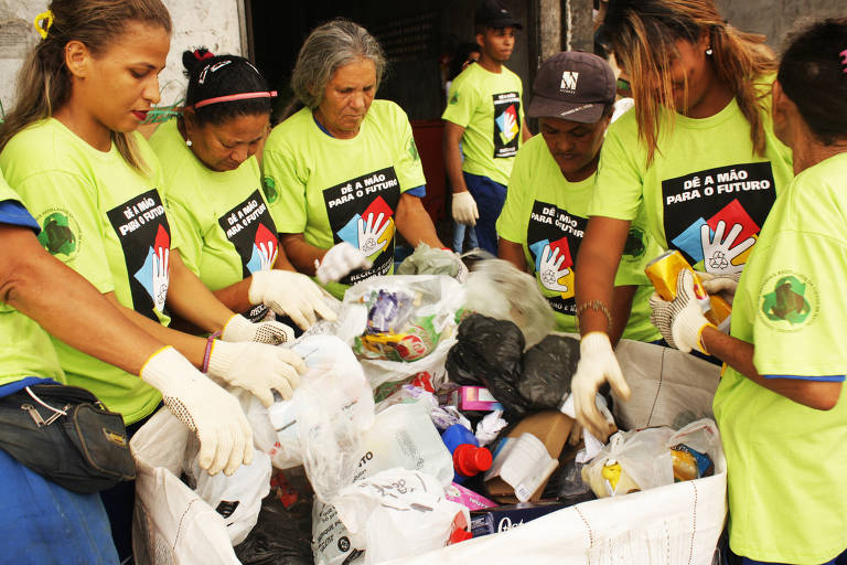 Mulheres catadoras com uniforme verde em torno de uma saco com produtos recicláveis