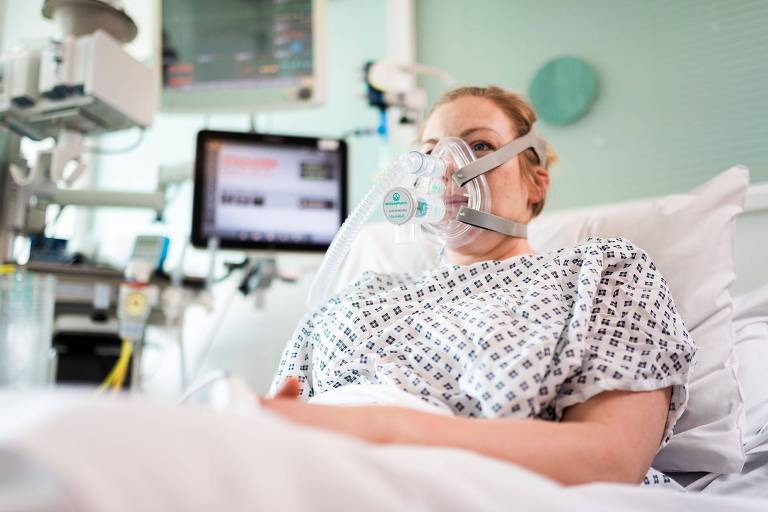 Mulher em leito de hospital, com camisola, usa o respirador no rosto 