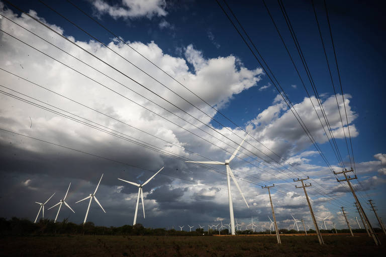 A imagem mostra um parque eólico com várias turbinas eólicas em um campo aberto. Ao fundo， há nuvens escuras e um céu azul. Linhas de transmissão de energia elétrica estão visíveis em primeiro plano， correndo paralelamente às turbinas.