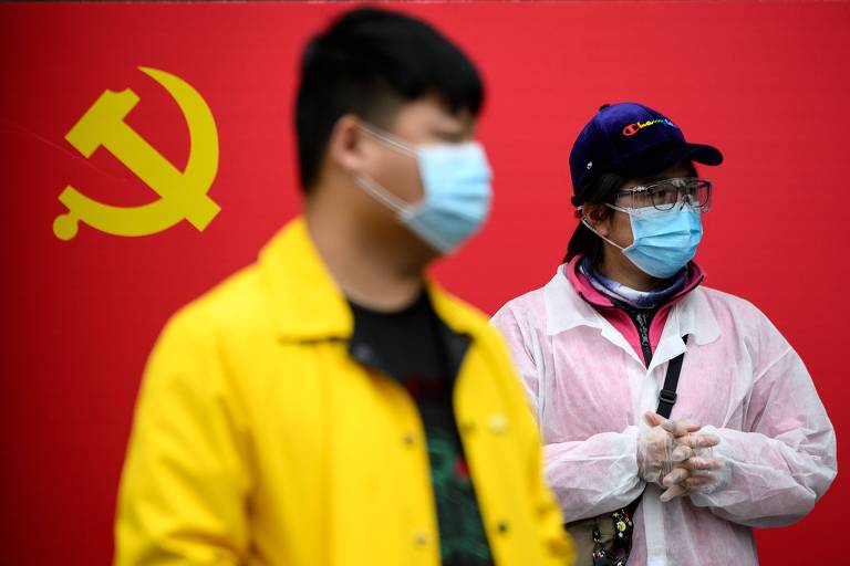 dois chineses vestindo máscaras olham para o lado direito; atrás deles, uma bandeira do partido comunista chinês
