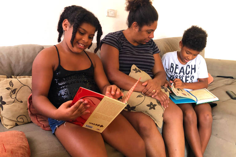  A pedagoga Simone Inácio, com os filhos, Isadora e Isaac, fazendo atividades em casa; estão os três sentados num sofá, a mãe entre a menina e o menino; as crianças folheiam livros