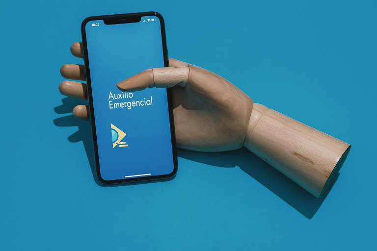 Uma foto com o fundo azul com uma mão articulada de madeira segurando um celular. Na tela, está aberto o aplicativo do auxílio emergencial.