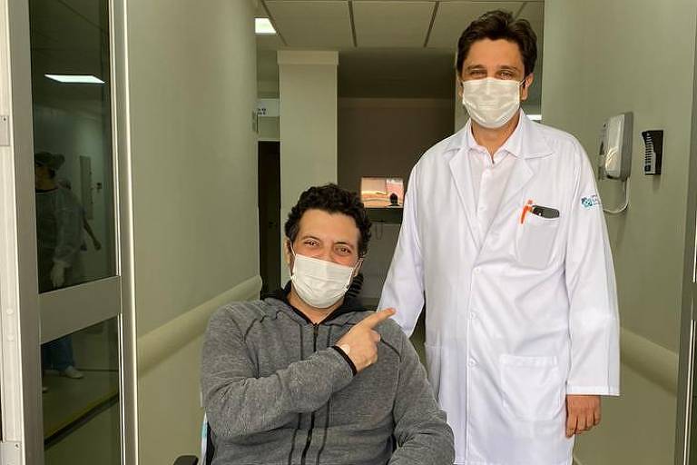 O paciente José Luiz, de casaco cinza, numa cadeira de rodas, aponta para o médico Marcelo, que está de pé ao seu lado, usando um jaleco branco; ambos usam máscara cirúrgica
