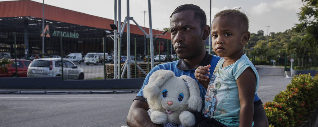 George Souza, 29, desempregado, com seu filho no colo, pede ajuda aos motoristas em frente a um supermercado na avenida Senador Teotônio Vilela, na zona sul de São Paulo 