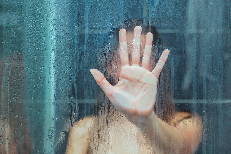 Pessoa colocando a mão no vidro de um box de banheiro, o que impede a visualização do rosto da pessoa