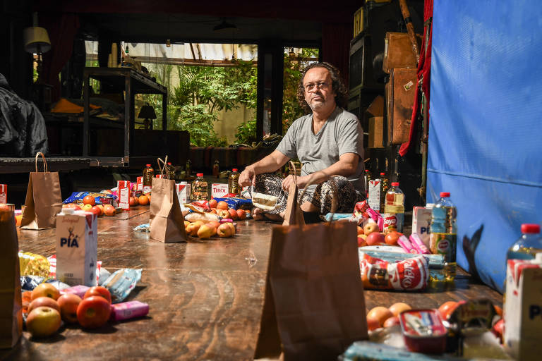 A imagem mostra um homem sentado no chão de um ambiente interno, cercado por sacolas de papel e diversos produtos alimentícios, como maçãs, caixas de suco e garrafas de óleo. O ambiente é iluminado e possui uma janela ao fundo, com vista para um espaço verde. O homem está usando uma camiseta cinza e calças estampadas, e parece estar organizando os itens ao seu redor.